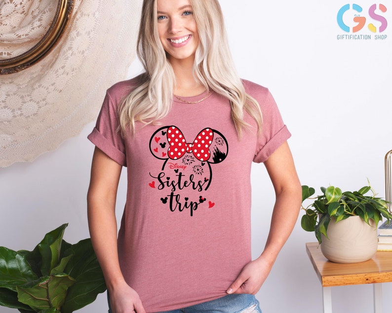 Puede incluir: Una camiseta rosa con un gr&aacute;fico en blanco y negro de orejas de Minnie Mouse con lunares rojos. El gr&aacute;fico tambi&eacute;n incluye el texto "Disney Sisters Trip" en negro.