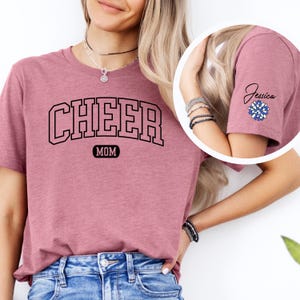 May include: A mauve t-shirt with the text "CHEER MOM" in black. The shirt has a blue and white pom-pom on the sleeve with the name "Jessica" written in black.