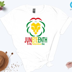 Pode incluir: Camiseta branca com um design colorido para o Juneteenth. O design inclui uma cabe&ccedil;a de le&atilde;o amarela, detalhes verdes e vermelhos e o texto "Juneteenth Is My Independence Day."