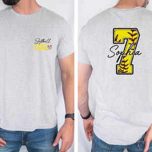 May include: A gray t-shirt with the text "Softball Dad" on the front and a yellow softball with the number 7 and the name "Sophia" on the back.