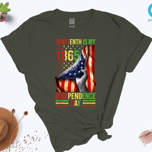 Puede incluir: Camiseta verde oliva oscuro con un dise&ntilde;o gr&aacute;fico. El dise&ntilde;o presenta el texto "Juneteenth is my 1865 Independence Day" junto con una bandera estadounidense en rojo, amarillo y verde. Una prenda casual.