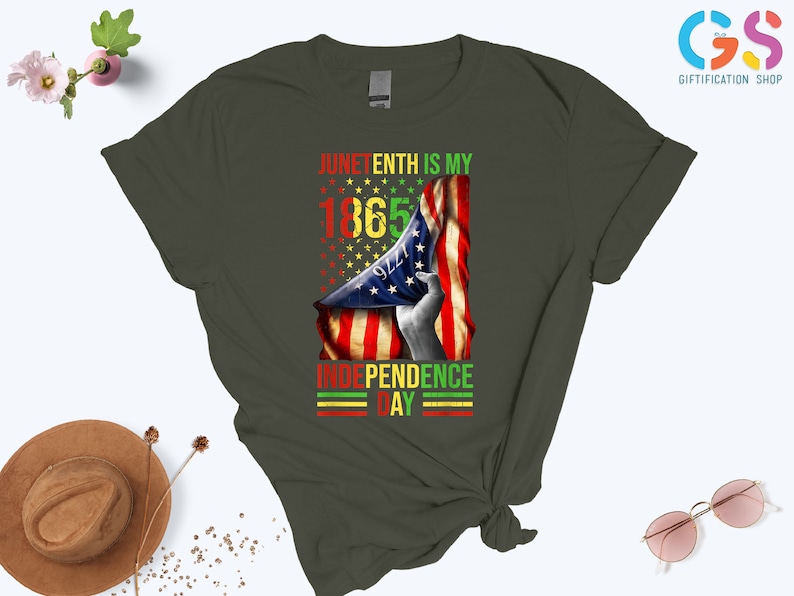 Puede incluir: Camiseta verde oliva oscuro con un dise&ntilde;o gr&aacute;fico. El dise&ntilde;o presenta el texto "Juneteenth is my 1865 Independence Day" junto con una bandera estadounidense en rojo, amarillo y verde. Una prenda casual.