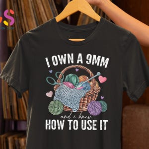 May include: Black t-shirt with white text that reads "I own a 9mm and I know how to use it". The shirt features a graphic of a basket filled with yarn and knitting needles.