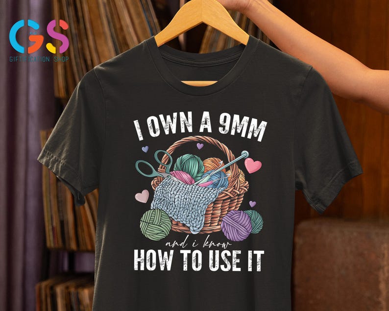 May include: Black t-shirt with white text that reads "I own a 9mm and I know how to use it". The shirt features a graphic of a basket filled with yarn and knitting needles.