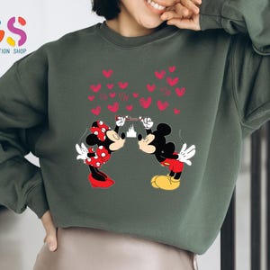 May include: Dark green sweatshirt featuring a graphic of Mickey and Minnie Mouse kissing, with red hearts and a castle in the background. Minnie wears a red polka dot bow and dress. Mickey wears red shorts and yellow shoes. The sweatshirt has the text "Giftification Shop".