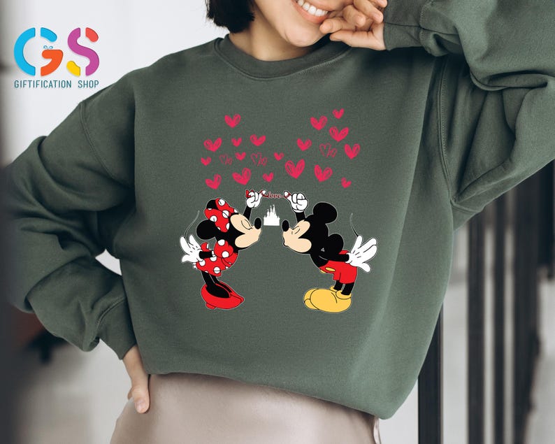 May include: Dark green sweatshirt featuring a graphic of Mickey and Minnie Mouse kissing, with red hearts and a castle in the background. Minnie wears a red polka dot bow and dress. Mickey wears red shorts and yellow shoes. The sweatshirt has the text "Giftification Shop".