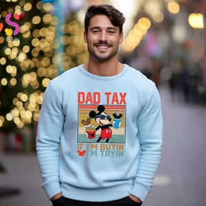 May include: Light blue sweatshirt featuring a graphic of Mickey Mouse wearing sunglasses, holding a drink and fries, with the text "DAD TAX" and "IF I'M BUYIN' I'M TRYIN'". The sweatshirt is a crew neck style.