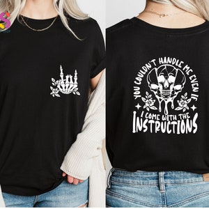 Op de afbeelding: Zwart T-shirt met een witte skelet hand die een rock and roll-teken maakt op de voorkant en de tekst "You couldn't handle me even if I come with the instructions" op de achterkant.