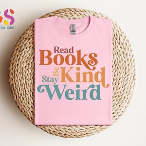 K&ouml;nnte beinhalten: Pinkes T-Shirt mit dem Text "Read Books Be Kind Stay Weird" in einer Retro-Schriftart mit orange, braun und t&uuml;rkisfarbenen Farben.