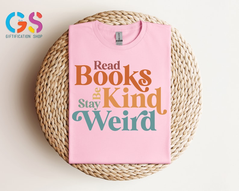 K&ouml;nnte beinhalten: Pinkes T-Shirt mit dem Text "Read Books Be Kind Stay Weird" in einer Retro-Schriftart mit orange, braun und t&uuml;rkisfarbenen Farben.