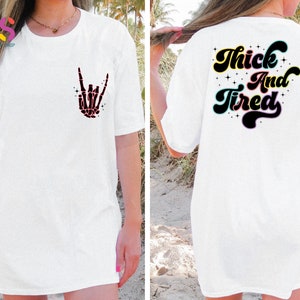 Op de afbeelding: Wit T-shirt met een skelethand die een rock-and-roll-teken maakt op de voorkant en de tekst "Thick And Tired" in een retro-lettertype op de achterkant.