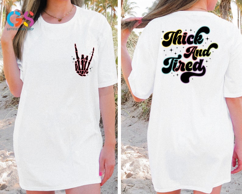 Op de afbeelding: Wit T-shirt met een skelethand die een rock-and-roll-teken maakt op de voorkant en de tekst "Thick And Tired" in een retro-lettertype op de achterkant.