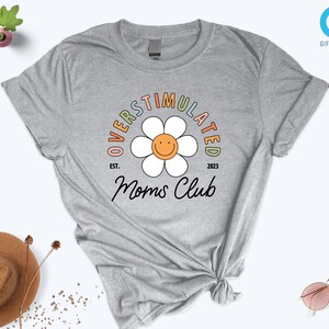May include: Gray t-shirt with a rainbow colored graphic of a daisy with a smiley face. The text "Overstimulated Moms Club" is written around the daisy with the text "Est. 2023" below the daisy.
