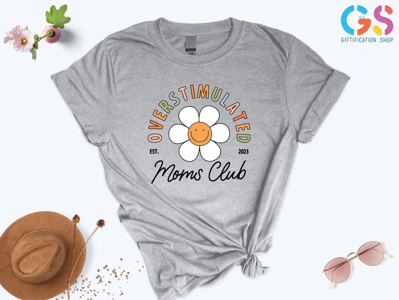 May include: Gray t-shirt with a rainbow colored graphic of a daisy with a smiley face. The text "Overstimulated Moms Club" is written around the daisy with the text "Est. 2023" below the daisy.