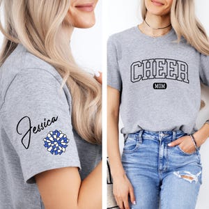 May include: Gray t-shirt with the text "CHEER MOM" in a black arched font. The shirt also features a blue and white pom-pom on the sleeve with the name "Jessica" written in cursive.