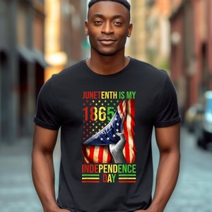 Puede incluir: Camiseta negra con el texto "Juneteenth is my 1865 Independence Day" en rojo, amarillo, verde y negro. El dise&ntilde;o incluye una mano sosteniendo una bandera estadounidense ondeando.