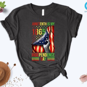 Puede incluir: Camiseta gris oscuro con un dise&ntilde;o gr&aacute;fico que celebra el Juneteenth. El dise&ntilde;o incluye el texto "Juneteenth is my 1865 Independence Day" con una bandera estadounidense y estrellas.