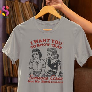 Op de afbeelding: Grijs T-shirt met een afbeelding van twee vrouwen en de tekst "Ik wil dat je weet dat iemand geeft, niet ik, maar iemand".