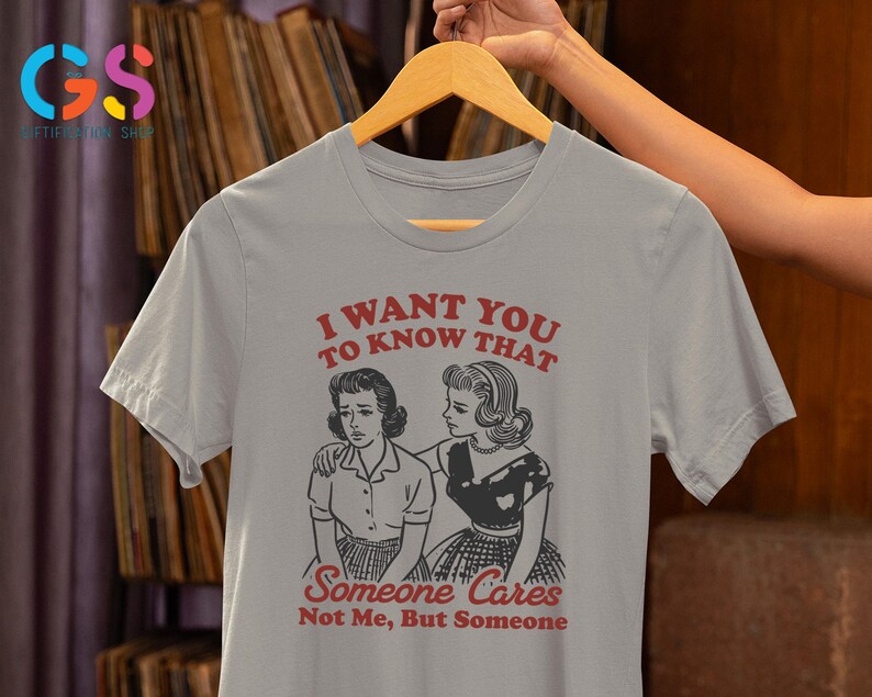 Op de afbeelding: Grijs T-shirt met een afbeelding van twee vrouwen en de tekst "Ik wil dat je weet dat iemand geeft, niet ik, maar iemand".