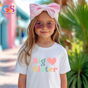 May include: A white t-shirt with the words "Big Sister" in colorful letters and a pink heart. The girl is wearing a pink headband and sunglasses.