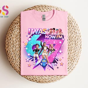 May include: Pink t-shirt with the text "I WAS 6, NOW I'M 7" and a graphic of three animated characters. The number 6 is teal glitter, and the number 7 is pink glitter. Stars and a blue cartoon character are also featured.