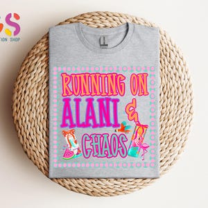 Puede incluir: Una camiseta gris jaspeado con el texto "RUNNING ON ALANI & CHAOS" en rosa y naranja. El dise&ntilde;o incluye ilustraciones de un flamenco, un loro y una rodaja de sand&iacute;a. La camiseta est&aacute; doblada sobre una superficie circular tejida.