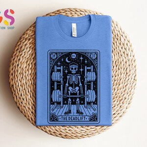 May include: A blue t-shirt featuring a black and white illustration of a skeleton lifting weights. The illustration is in the style of a tarot card and includes the text "The Deadlift".