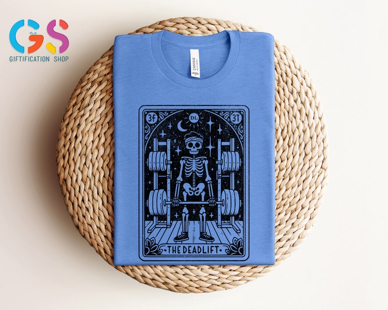 May include: A blue t-shirt featuring a black and white illustration of a skeleton lifting weights. The illustration is in the style of a tarot card and includes the text "The Deadlift".