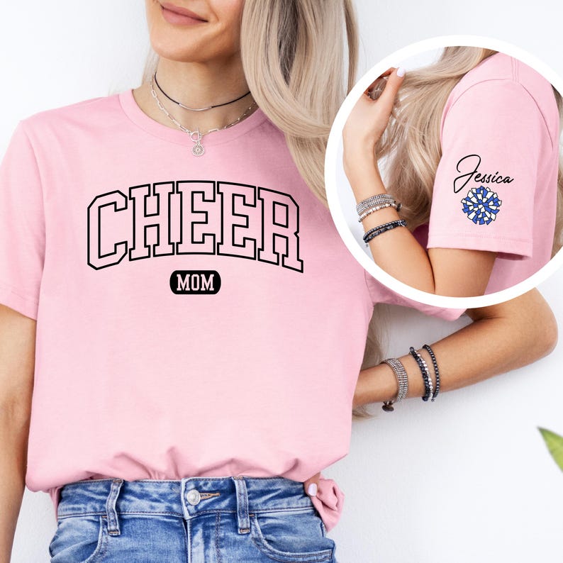 May include: Pink t-shirt with the text "CHEER MOM" in black. The shirt has a blue and white pom-pom on the sleeve with the name "Jessica" written above it.