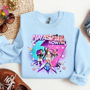 以下が含まれることがあります： グラフィックデザインが施された水色のスウェットシャツ。「I WAS 6 NOW I'M 7」の文字、人物のイラスト、星が描かれています。スウェットシャツは柔らかい素材で作られています。デザインは青とピンクの配色を使用しています。