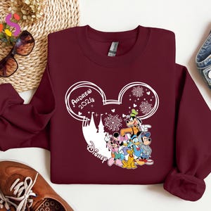Pode incluir: Um moletom bord&ocirc; com um design branco da cabe&ccedil;a do Mickey Mouse, o texto "Andrew 2026" e uma silhueta do castelo da Disney. O design inclui personagens da Disney e fogos de artif&iacute;cio.