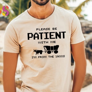 Puede incluir: Camiseta beige con texto negro que dice "Por favor, ten paciencia conmigo, soy de los a&ntilde;os 1900". El texto est&aacute; en una fuente pixelada. Debajo del texto hay una imagen en blanco y negro de un carro tirado por un buey.