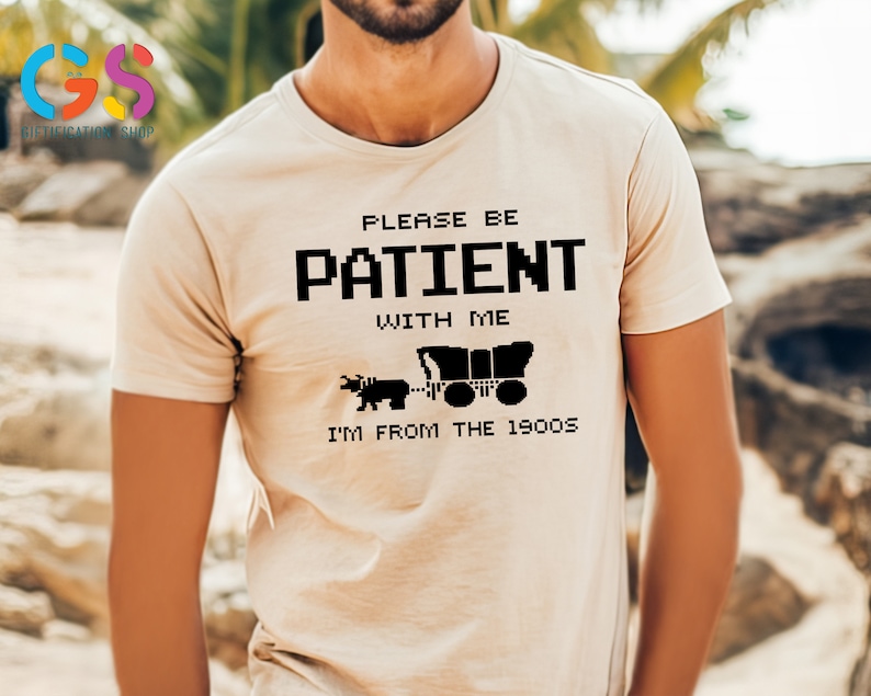 Puede incluir: Camiseta beige con texto negro que dice "Por favor, ten paciencia conmigo, soy de los a&ntilde;os 1900". El texto est&aacute; en una fuente pixelada. Debajo del texto hay una imagen en blanco y negro de un carro tirado por un buey.