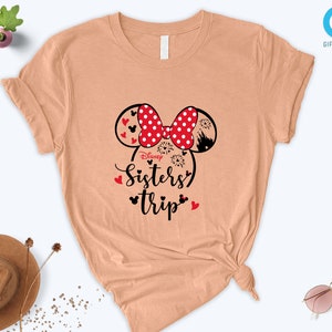 Puede incluir: Una camiseta de color melocot&oacute;n con un gr&aacute;fico en blanco y negro de orejas de Mickey Mouse con lazos de lunares rojos. El texto "Disney Sisters Trip" est&aacute; impreso debajo de las orejas.