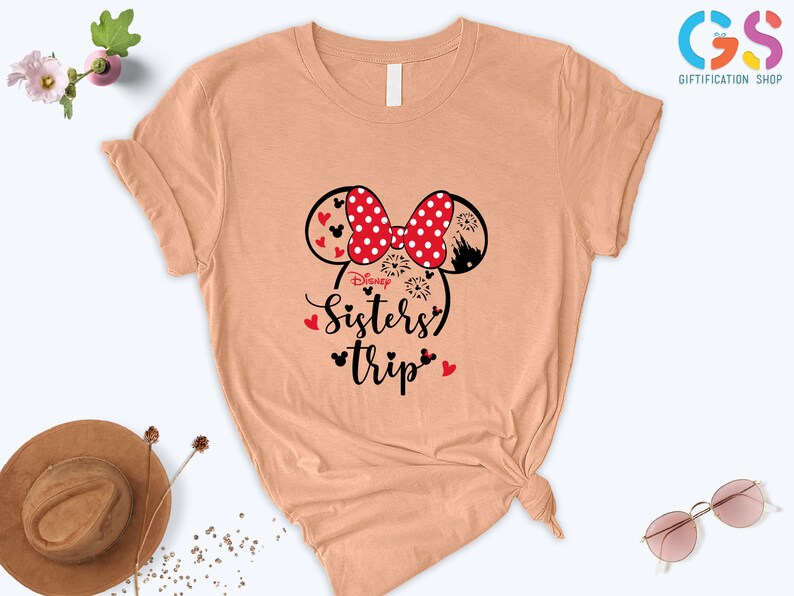Puede incluir: Una camiseta de color melocot&oacute;n con un gr&aacute;fico en blanco y negro de orejas de Mickey Mouse con lazos de lunares rojos. El texto "Disney Sisters Trip" est&aacute; impreso debajo de las orejas.