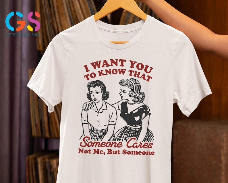 Op de afbeelding: Wit T-shirt met een zwart-witte illustratie van twee vrouwen. De tekst op het shirt luidt "Ik wil dat je weet dat iemand geeft, niet ik, maar iemand."