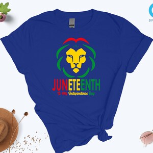 Pode incluir: Uma camiseta azul royal com um desenho de cabe&ccedil;a de le&atilde;o em amarelo, verde e vermelho. O texto "JUNETEENTH" &eacute; exibido abaixo do le&atilde;o, com "Is My Independence Day" abaixo. A camisa tem um n&oacute; amarrado na parte inferior.