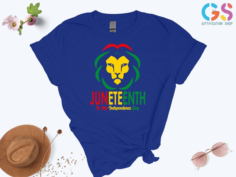 Pode incluir: Uma camiseta azul royal com um desenho de cabe&ccedil;a de le&atilde;o em amarelo, verde e vermelho. O texto "JUNETEENTH" &eacute; exibido abaixo do le&atilde;o, com "Is My Independence Day" abaixo. A camisa tem um n&oacute; amarrado na parte inferior.