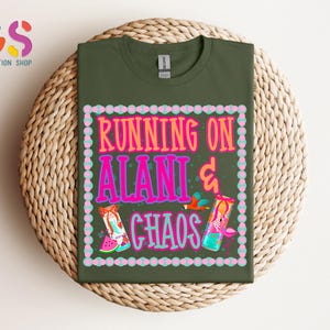 Puede incluir: Una camiseta verde oliva con el texto "RUNNING ON ALANI & CHAOS" en rosa y naranja, con ilustraciones coloridas. La camiseta se muestra sobre una superficie circular tejida.