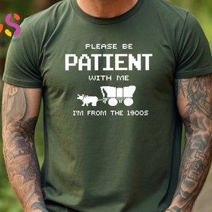 Puede incluir: Una camiseta verde con texto blanco que dice "Por favor, ten paciencia conmigo, soy de los a&ntilde;os 1900". El texto est&aacute; en una fuente pixelada y hay una imagen pixelada de un carro cubierto tirado por un buey.