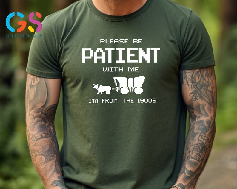 Puede incluir: Una camiseta verde con texto blanco que dice "Por favor, ten paciencia conmigo, soy de los a&ntilde;os 1900". El texto est&aacute; en una fuente pixelada y hay una imagen pixelada de un carro cubierto tirado por un buey.