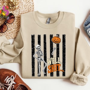 May include: A beige crewneck sweatshirt featuring a graphic of a skeleton holding a string attached to an orange jack-o'-lantern balloon. The design includes vertical black distressed stripes and the orange text "WELL, SHIT..." in a bold font.