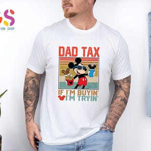 May include: White t-shirt featuring a graphic of Mickey Mouse wearing sunglasses and holding food and a drink. The text "DAD TAX" is above the graphic, with "IF I'M BUYIN' I'M TRYIN'" below. The shirt is a casual wear item.