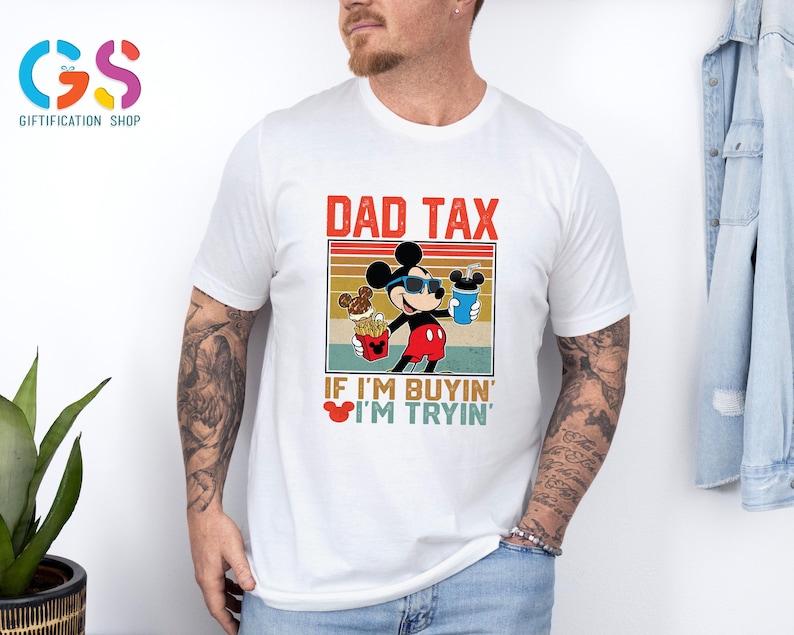 May include: White t-shirt featuring a graphic of Mickey Mouse wearing sunglasses and holding food and a drink. The text "DAD TAX" is above the graphic, with "IF I'M BUYIN' I'M TRYIN'" below. The shirt is a casual wear item.