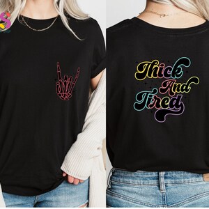 Op de afbeelding: Zwart T-shirt met een roze skelet hand die een rock and roll-teken maakt op de voorkant en de tekst "Thick And Tired" in regenboogkleuren op de achterkant.