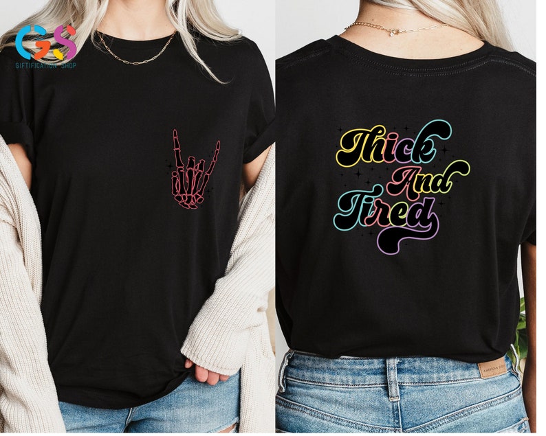 Op de afbeelding: Zwart T-shirt met een roze skelet hand die een rock and roll-teken maakt op de voorkant en de tekst "Thick And Tired" in regenboogkleuren op de achterkant.
