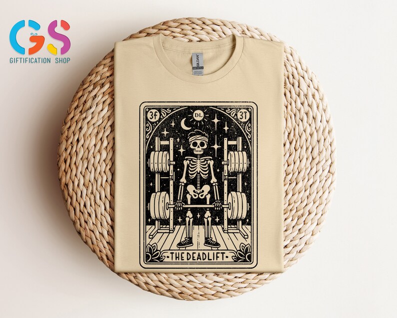 May include: A beige t-shirt featuring a black and white illustration of a skeleton lifting weights. The illustration is framed in a black border with the text "The Deadlift" at the bottom.