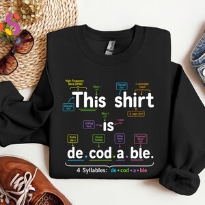 May include: A black long-sleeve sweatshirt featuring the white text "This shirt is de.cod.a.ble." with colorful linguistic annotations and arrows explaining phonics concepts like digraphs, syllables, and prefixes printed across the front.