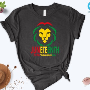 Pode incluir: T-shirt cinzenta escura com um desenho de cabe&ccedil;a de le&atilde;o em amarelo, verde e vermelho, e a palavra "JUNETEENTH" em vermelho e verde. O texto "is my Independence Day" est&aacute; abaixo. Uma pe&ccedil;a de roupa casual.