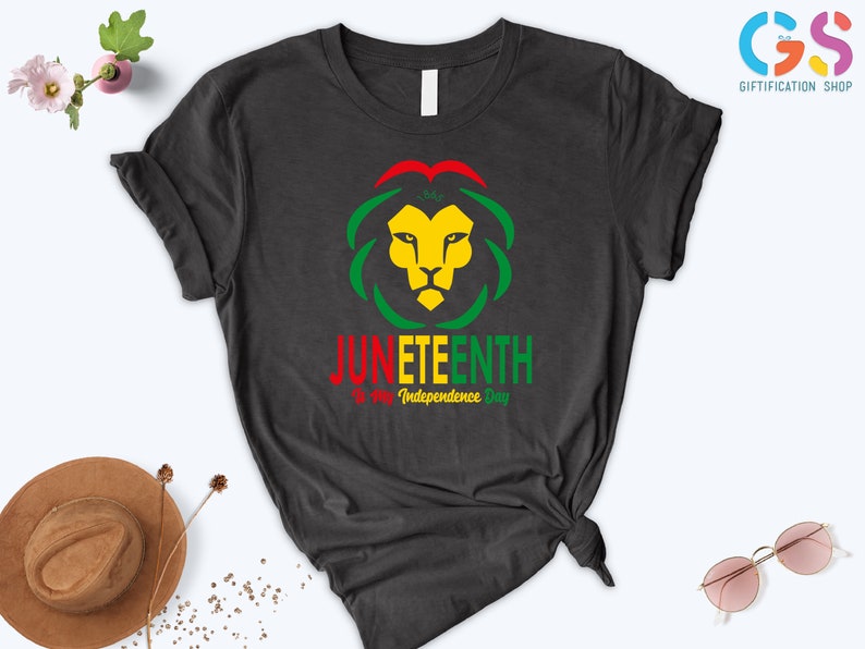 Pode incluir: T-shirt cinzenta escura com um desenho de cabe&ccedil;a de le&atilde;o em amarelo, verde e vermelho, e a palavra "JUNETEENTH" em vermelho e verde. O texto "is my Independence Day" est&aacute; abaixo. Uma pe&ccedil;a de roupa casual.